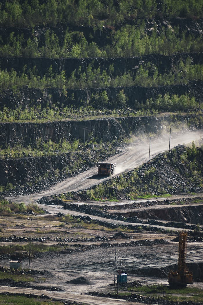 A truck kicks up dust in a sprawling quarry surrounded by green foliage.